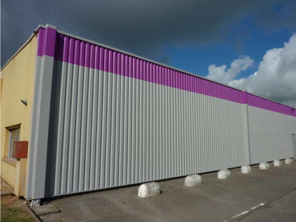 Długi budynek przemysłowy z szarą, falistą elewacją i fioletowym pasem u góry, widoczny z zewnątrz w pochmurny dzień.