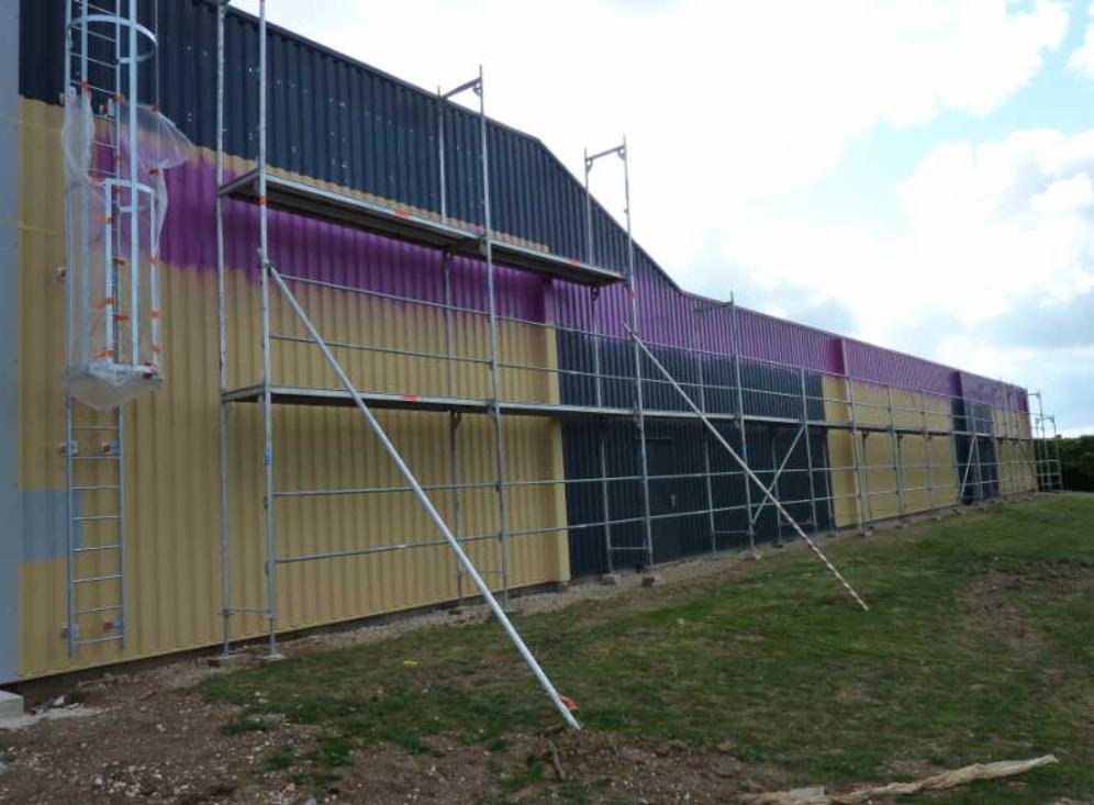 Malowanie elewacji budynku przemysłowego na żółto, fioletowo i grafitowo z użyciem rusztowań, widoczne początki malowania fioletem.