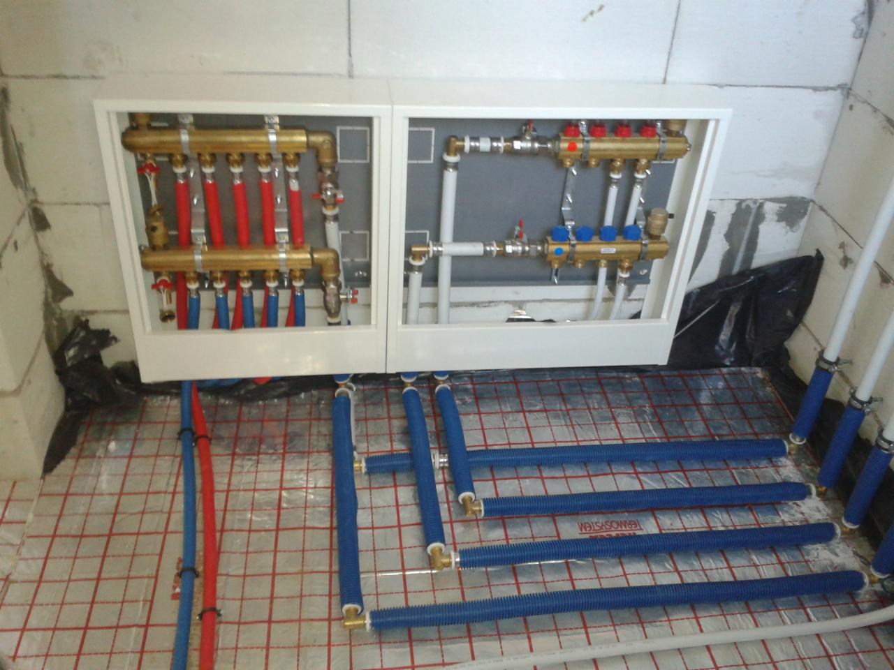 Rozdzielacz ogrzewania podłogowego w białej obudowie, z widocznymi rurami w izolacji na folii z rastrem, w surowym wnętrzu. Detal instalacji.