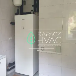 Biała jednostka wewnętrzna z zbiornikiem buforowym, rury w izolacji, logo firmy Rapacz HVAC na tle jasnych płytek ceramicznych.