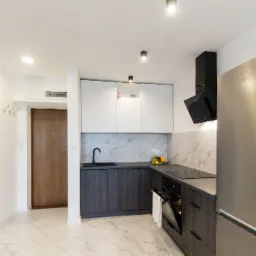 Nowoczesna kuchnia z szarymi szafkami dolnymi i białymi górnymi, marmurowym backsplash'em, stalową lodówką i czarną armaturą, widok na przedpokój z lustrem i drzwiami.