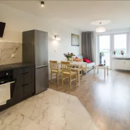 Widok na otwartą przestrzeń mieszkania po remoncie: kuchnia z marmurową podłogą przechodząca w drewnianą podłogę salonu, stół z krzesłami, sofa i balkon z widokiem na zewnątrz.