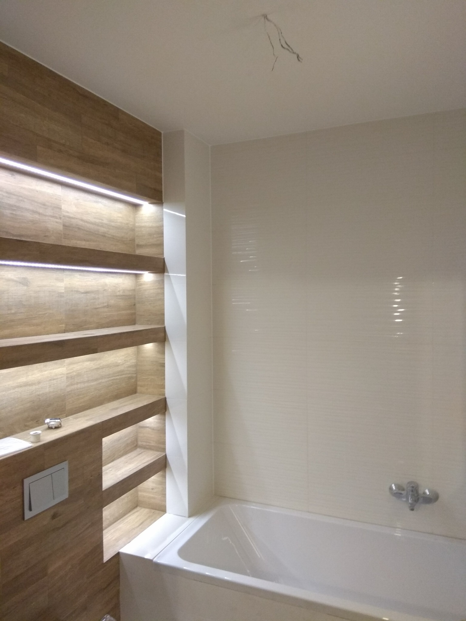 Nowoczesna łazienka z podświetlanymi półkami z płytek imitujących drewno, białą wanną i minimalistyczną armaturą.