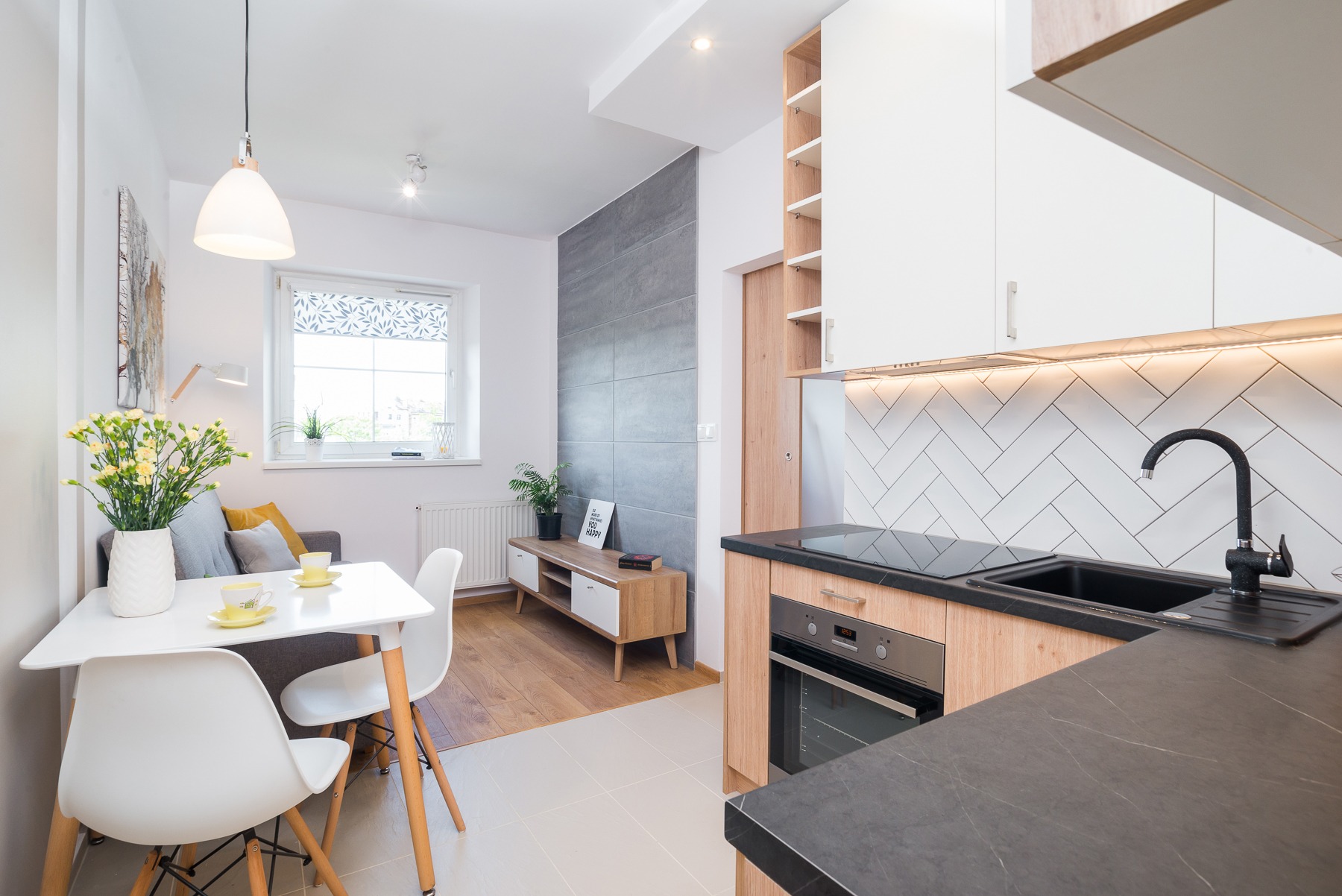 Jasne wnętrze mieszkania z aneksem kuchennym, białymi szafkami, czarnym zlewem i blatem, oraz stołem z kwiatami i widokiem na salon z szarą sofą.