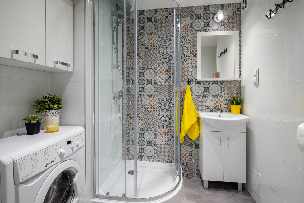 Nowoczesna łazienka w jasnych kolorach z kabiną prysznicową, umywalką z szafką, pralką i dekoracyjnymi płytkami na ścianie.