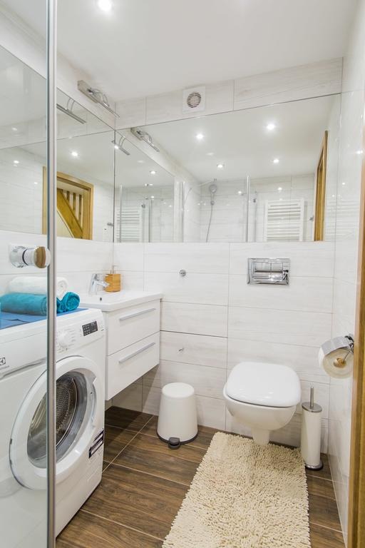 Jasna łazienka z pralką Electrolux, umywalką z szafką, toaletą podwieszaną i dużym lustrem na ścianie z białymi płytkami imitującymi drewno.