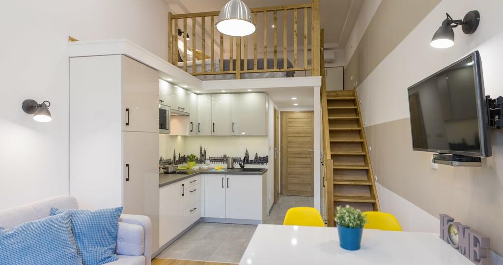 Wnętrze mieszkania z antresolą sypialnianą nad kuchnią, białe meble kuchenne z czarnymi uchwytami, schody drewniane, telewizor na ścianie, sofa z niebieskimi poduszkami, żółte krzesła przy białym...