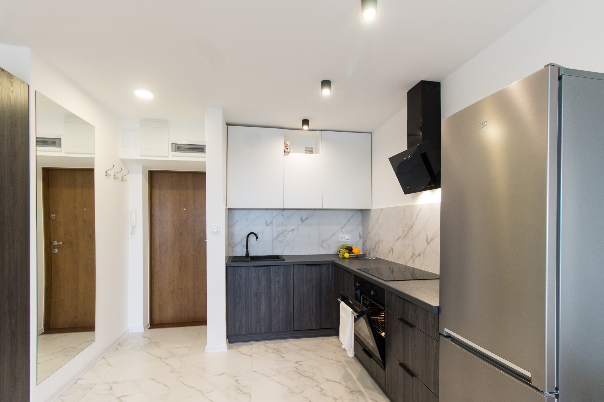 Nowoczesna kuchnia z szarymi szafkami dolnymi i białymi górnymi, marmurowym backsplash'em, stalową lodówką i czarną armaturą, widok na przedpokój z lustrem i drzwiami.