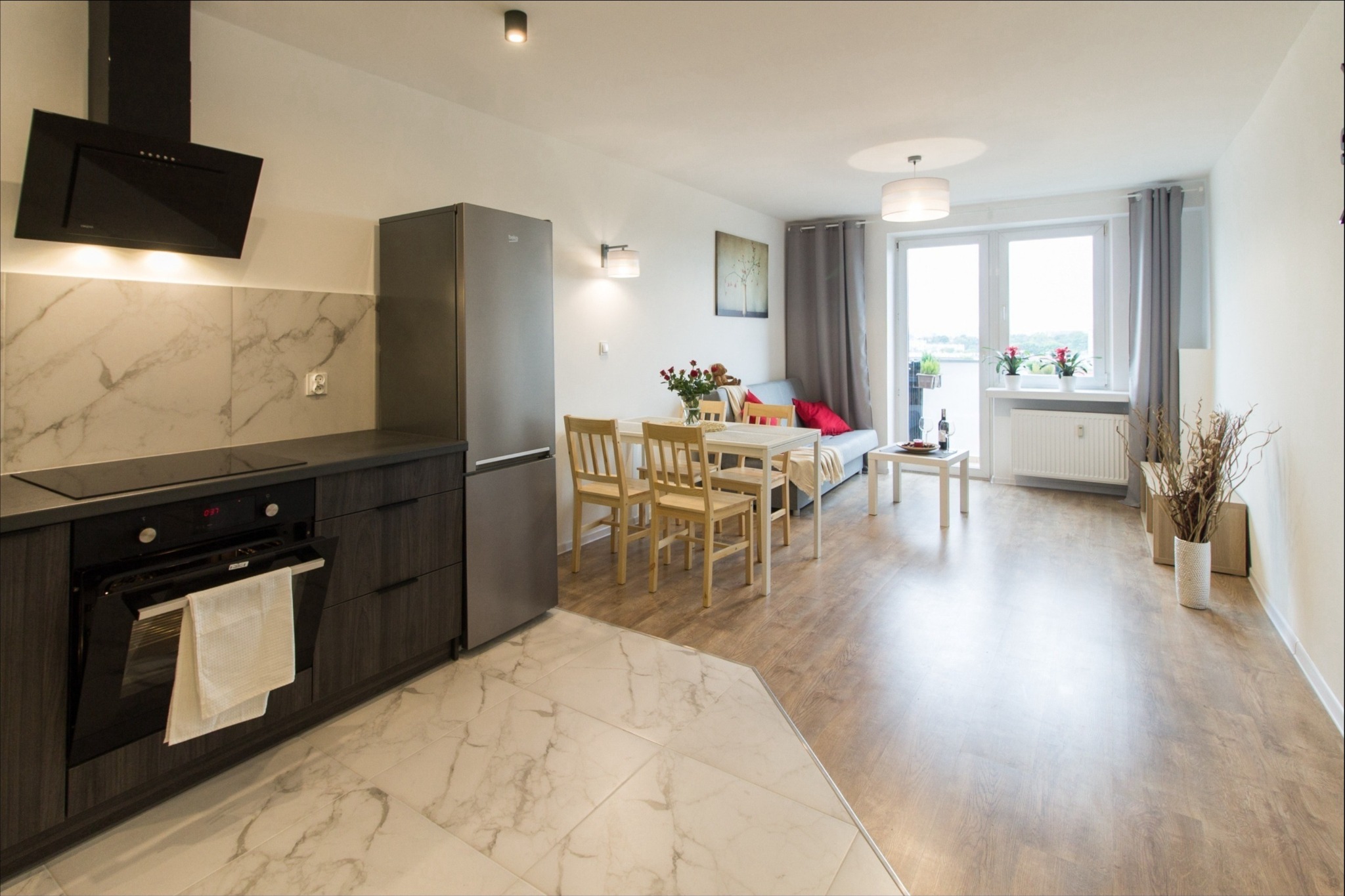 Widok na otwartą przestrzeń mieszkania po remoncie: kuchnia z marmurową podłogą przechodząca w drewnianą podłogę salonu, stół z krzesłami, sofa i balkon z widokiem na zewnątrz.