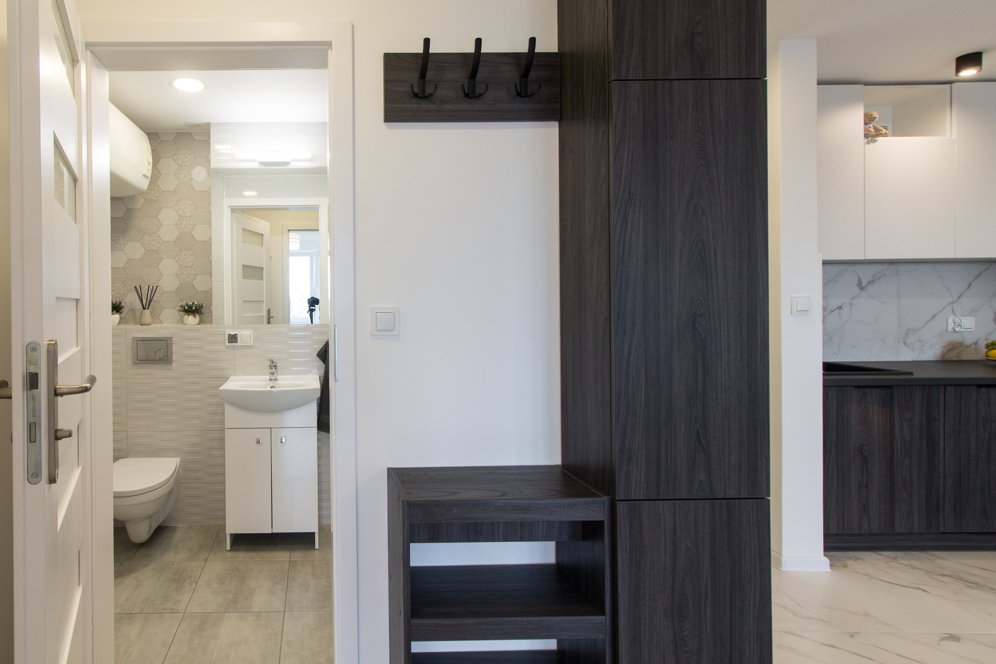 Nowoczesne wnętrze mieszkania z widokiem na łazienkę, szafa z wieszakami w przedpokoju i fragment kuchni w tle.