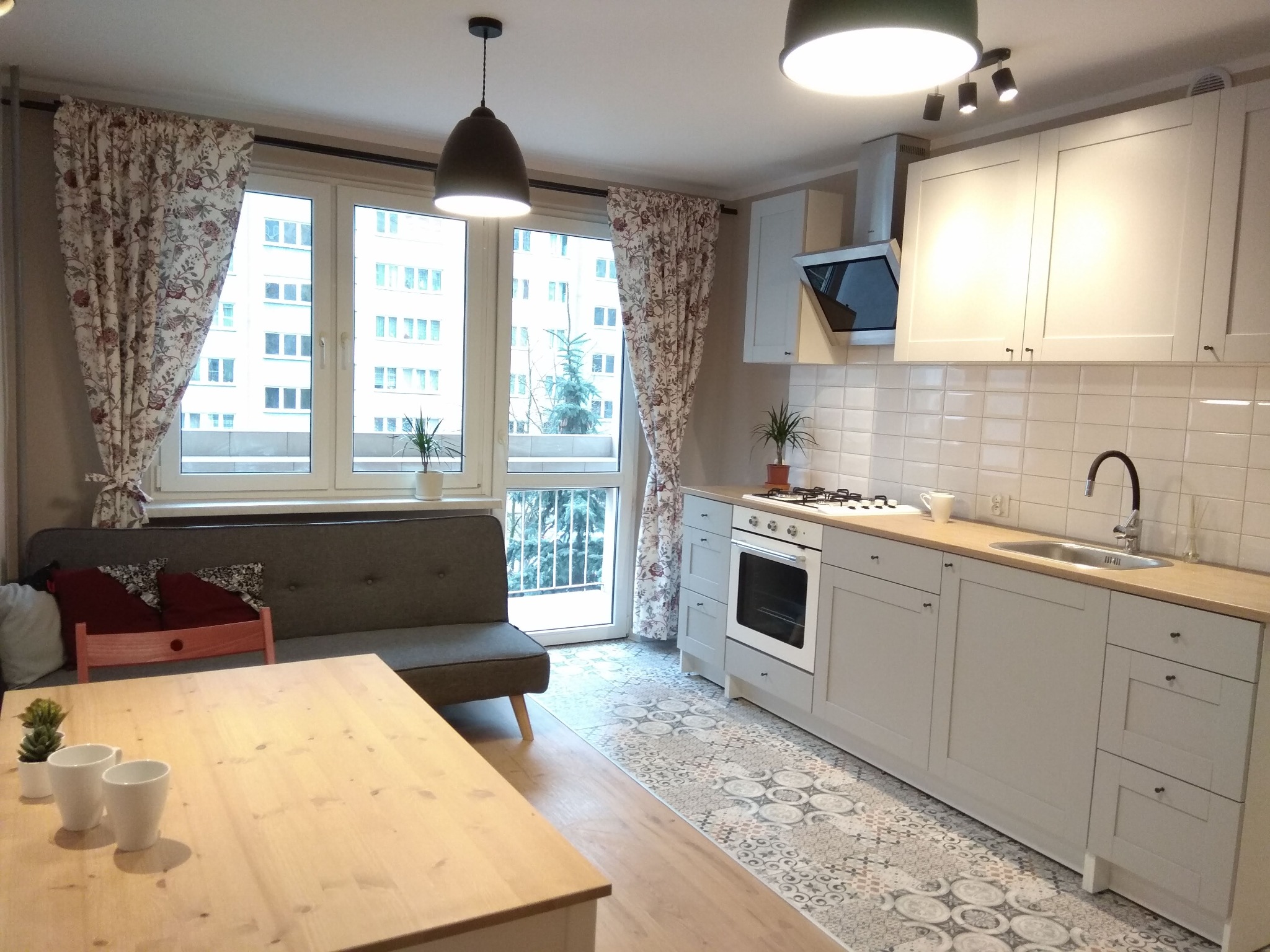 Jasne wnętrze po renowacji: kuchnia z szarymi szafkami, drewnianym blatem, wzorzystą podłogą i widokiem z okna na blok mieszkalny.