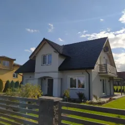 Nowoczesny dom murowany z ciemnym dachem i balkonem, widoczny zza metalowego ogrodzenia w słoneczny dzień.