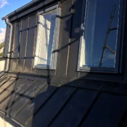 Czarny dach z blachy trapezowej z oknami dachowymi, widoczna rynna i drabina odbijająca się w szybie okna.