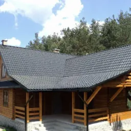 Gotowy dom z bali, z ciemnoszarą dachówką i drewnianą balustradą werandy, na tle lasu i błękitnego nieba z chmurami.