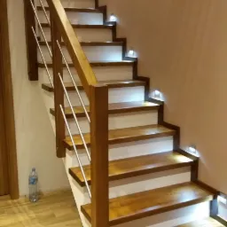 Drewniane schody z białymi podstopnicami, oświetleniem LED w stopniach i balustradą z drewnianymi słupkami i metalowymi prętami, widok z dołu schodów.