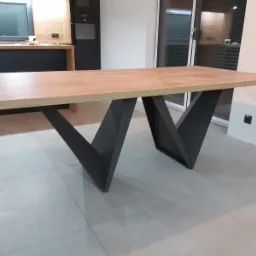 Stół z blatem z jasnego drewna i czarnymi, geometrycznymi nogami w kształcie litery V, na tle kuchni z zabudową w kolorach drewna i czerni.