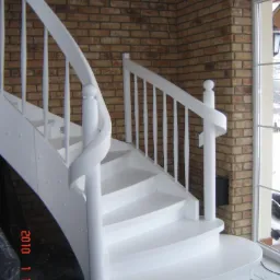 Białe, kręte schody drewniane z balustradą na tle ściany z cegły i okna.