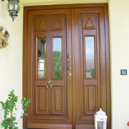 Dwuskrzydłowe wejściowe z przeszkleniami i lwem jako kołatką, widok z zewnątrz, na wycieraczce leżą klapki, obok doniczka z zieloną rośliną i lampion.