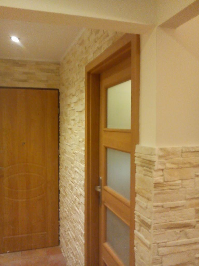 Korytarz z dekoracyjną ścianą z kamienia elewacyjnego w odcieniach beżu, sąsiadującą z drewnianą futryną i drzwiami z matowego szkła oraz brązowymi drzwiami wejściowymi.