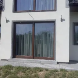 Biały budynek z brązowymi oknami i przesuwnymi drzwiami balkonowymi. Widoczne firanki w oknach i elementy wykończenia z szarego kamienia.