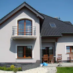 Dom z balkonem z balustradą ze stali nierdzewnej. Na balkonie widać drewniane okno z łukiem na górze. Pod balkonem okno z drewnianą ramą. Dach pokryty ciemną dachówką.