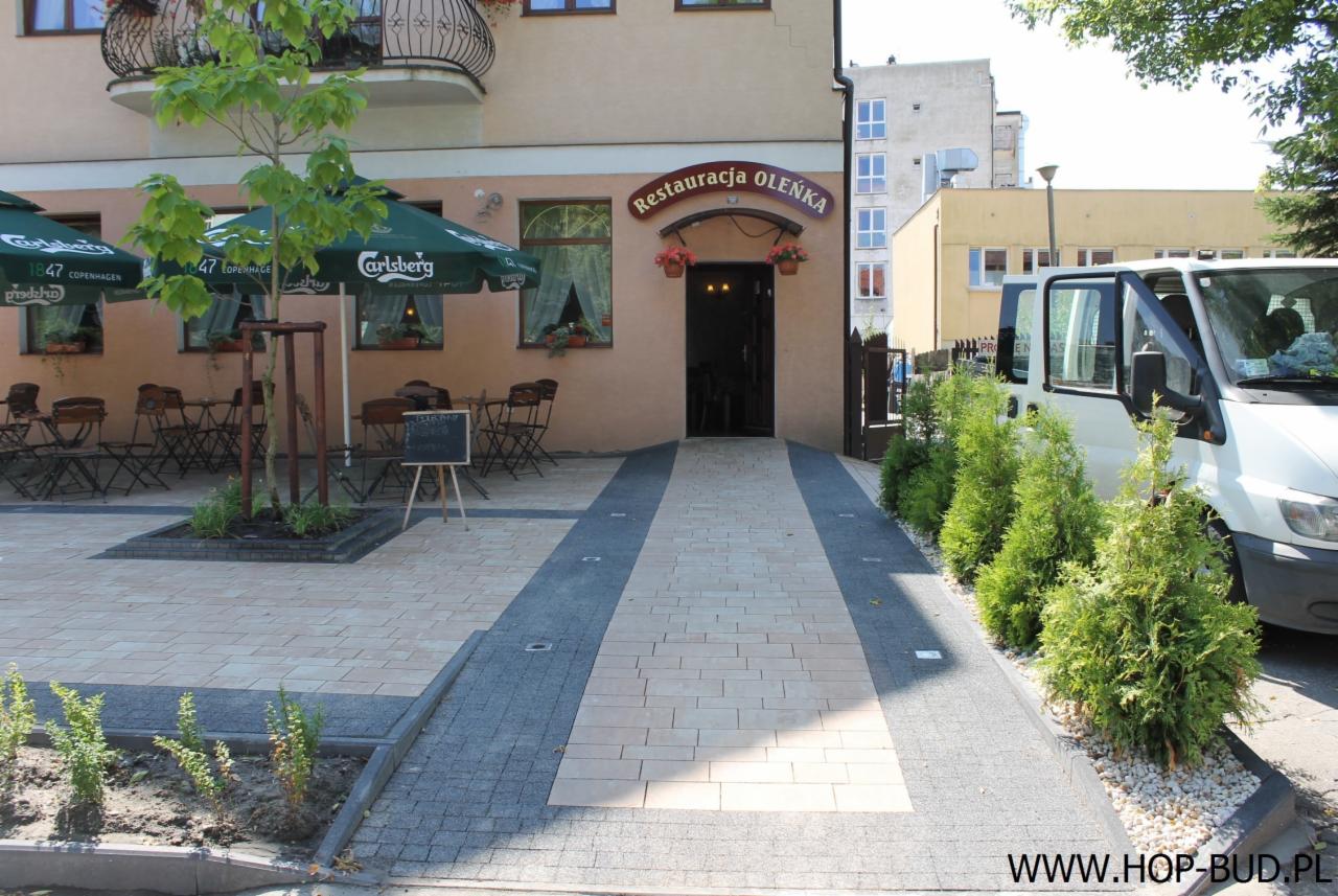 Ułożona kostka brukowa przed restauracją Oleńka, z rampą i roślinami. Widoczny van z otwartymi drzwiami, parasole z logo Carlsberg i tablica z menu.