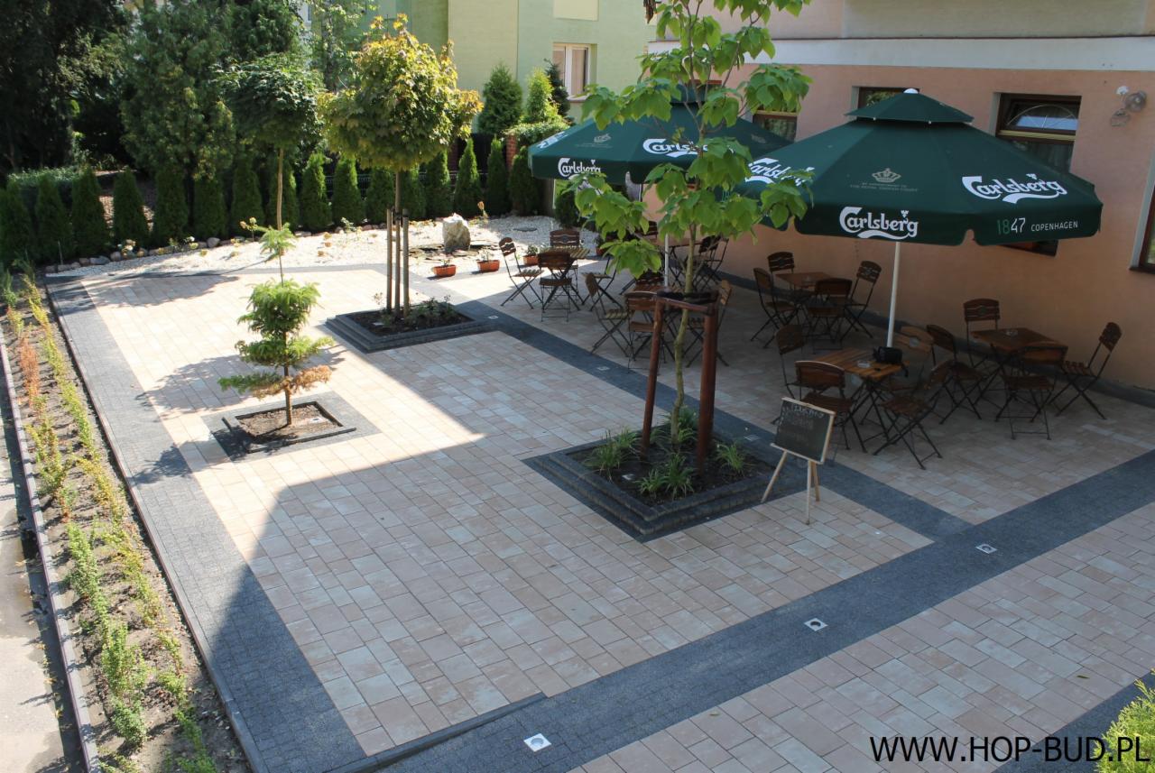 Ułożona kostka brukowa w patio z nasadzeniami drzew i krzewów, z parasolami i stolikami kawiarnianymi w tle. Widok z góry, słoneczny dzień.
