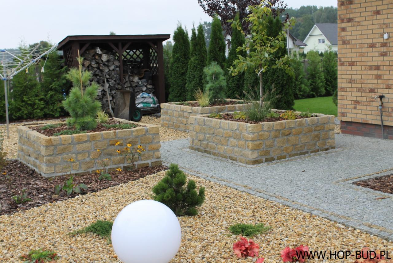 Ogród z kamiennymi rabatami, ścieżką z kostki i kulistą lampą. Rabaty w formie prostopadłościanów z roślinami. W tle drewutnia i tuje.