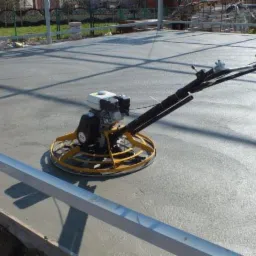 Zacieraczka mechaniczna w trakcie wygładzania świeżo wylanej, szarej posadzki jastrychowej na budowie. Widoczne metalowe profile i zarys konstrukcji budynku.