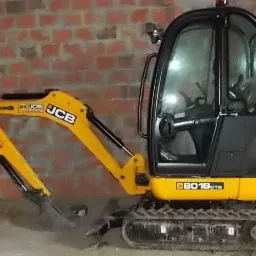 Żółta mini koparka JCB 8018 CTS z kabiną na gąsienicach, zaparkowana na szarym podłożu przed ścianą z czerwonej cegły.