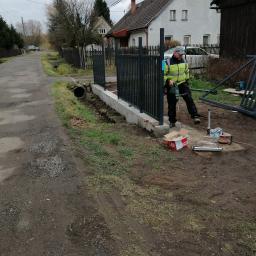 Seweryn - Pracownik w żółtej kurtce roboczej wierci otwory w betonowej podstawie pod metalowe przęsła ogrodzenia, widoczny fragment ulicy i domy w tle.