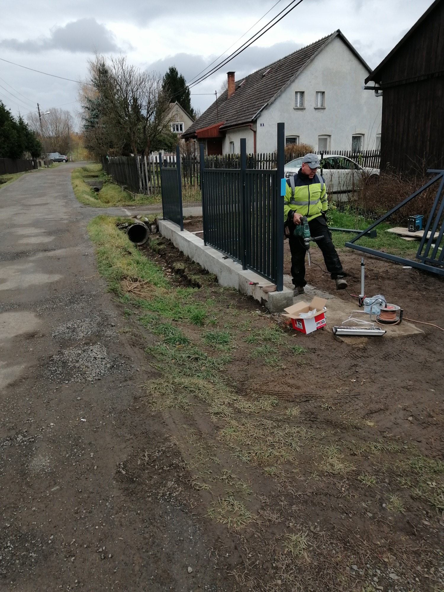 Pracownik w żółtej kurtce roboczej wierci otwory w betonowej podstawie pod metalowe przęsła ogrodzenia, widoczny fragment ulicy i domy w tle.