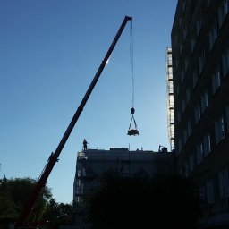 TopInstal Mariusz Wiśniewski - Montaż ciężkiego elementu na dachu budynku za pomocą dźwigu, sylwetka pracownika na dachu, w tle błękitne niebo i fragment budynku z oknami.