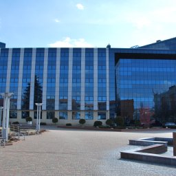 TopInstal Mariusz Wiśniewski - Nowoczesny budynek biurowy z przeszkloną fasadą odbijającą niebo, otoczony brukowanym placem z ławkami i oświetleniem.