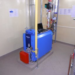 TopInstal Mariusz Wiśniewski - Niebieski piec Buderus z podłączonym laptopem, widoczne rury instalacji grzewczej w izolacji, czerwona obudowa palnika, żółta rura doprowadzająca gaz, dokumentacja techniczna na ścianie.