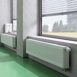 TopInstal Mariusz Wiśniewski - Dwa białe grzejniki panelowe zamontowane pod oknami na tle zielonej podłogi i surowych, betonowych ścian.