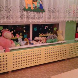 TopInstal Mariusz Wiśniewski - Żółta osłona grzejnika z perforacją w przedszkolu, za nią pluszaki na parapecie, okno z rysunkami i firanka z motywem kwiatowym.