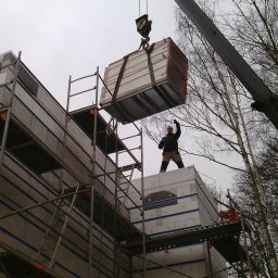 TopInstal Mariusz Wiśniewski - Montaż prefabrykowanych elementów budynku na placu budowy, z użyciem dźwigu i rusztowań, pracownik w kasku nadzoruje proces.