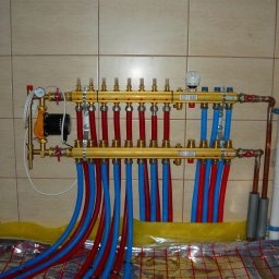 TopInstal Mariusz Wiśniewski - Rozdzielacz podłogowy z czerwonymi i niebieskimi rurami, zamontowany na ścianie z płytek, widoczne elementy instalacji i mata grzewcza pod folią