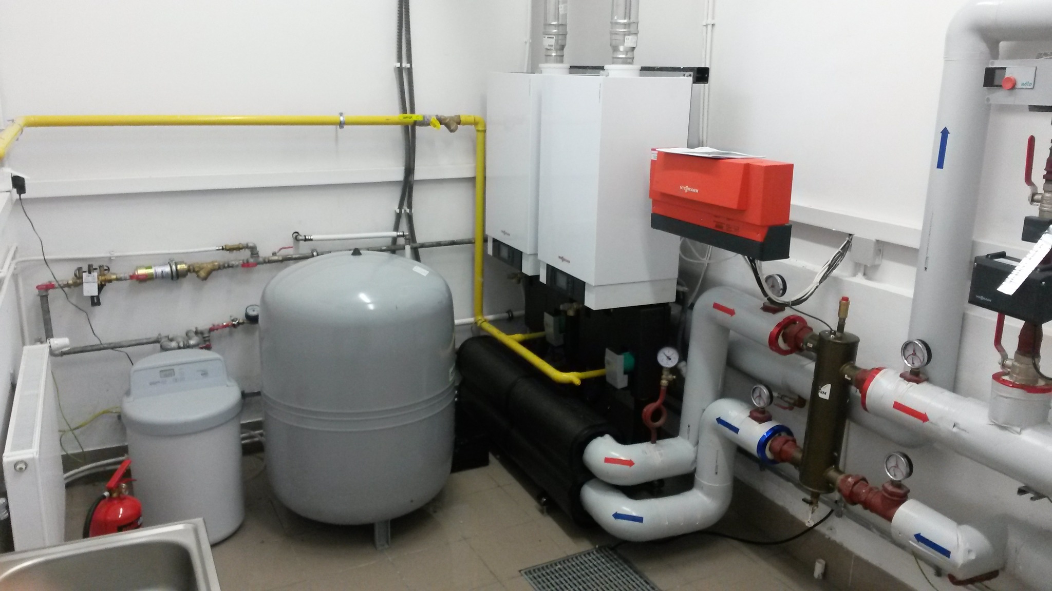 Kompletna kotłownia z dwoma białymi kotłami Viessmann, zbiornikiem wyrównawczym, zmiękczaczem wody i rurami oznaczonymi strzałkami przepływu, w pomieszczeniu z żółtą instalacją gazową.