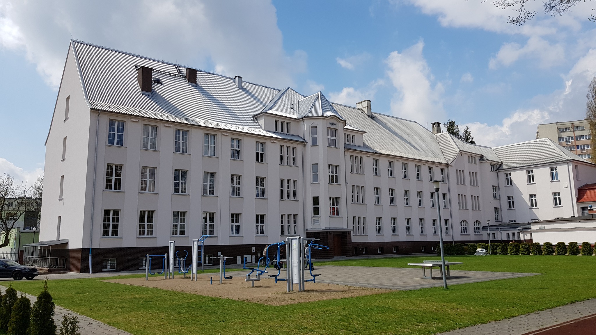 Duży, trzypiętrowy budynek z białej cegły z metalowym dachem, widoczny z zewnątrz, z placem zabaw na pierwszym planie, w słoneczny dzień.