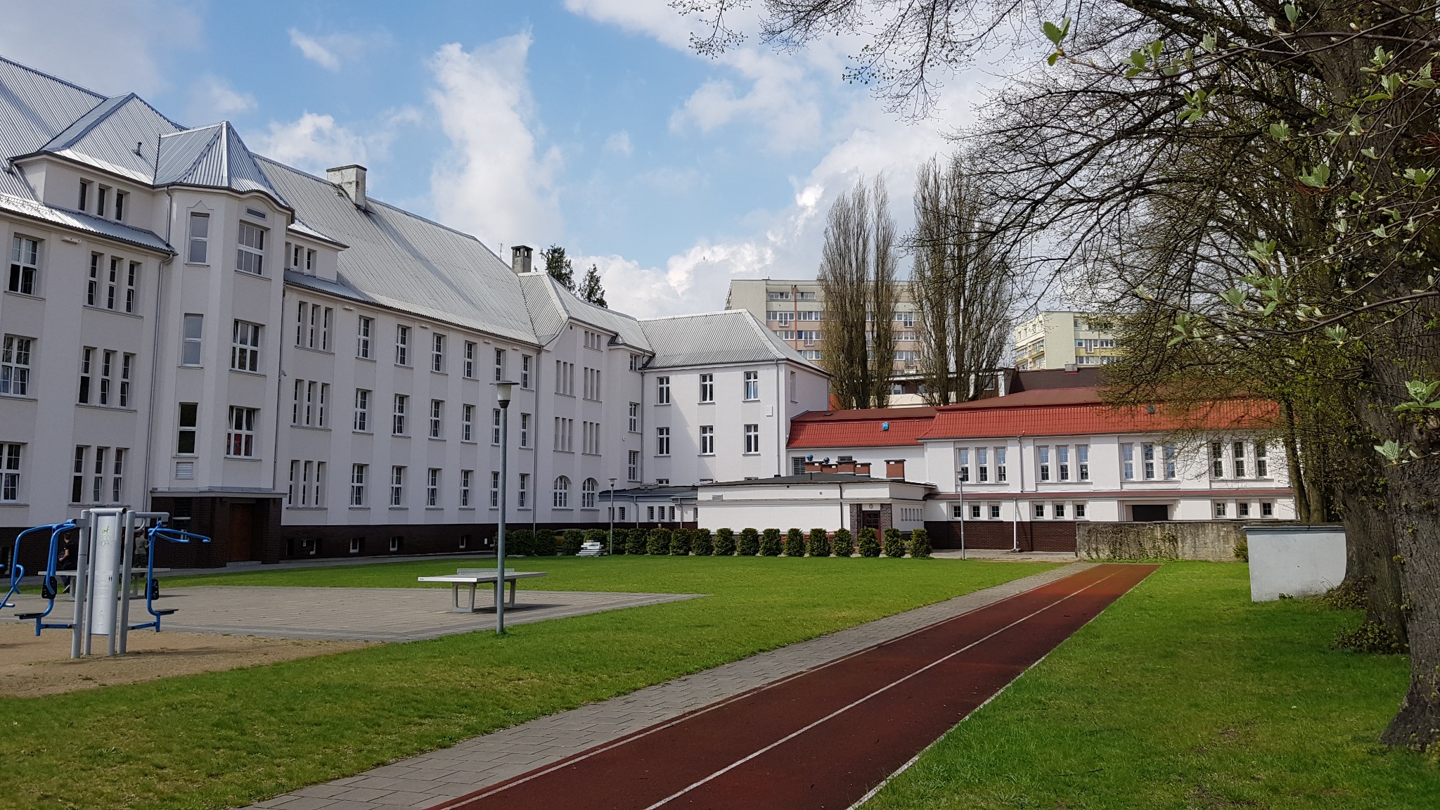 Budynek szkoły z szarym dachem, czerwona bieżnia i plac zabaw na trawniku w słoneczny dzień.