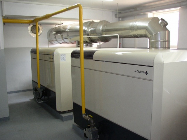 Dwa białe kotły grzewcze marki De Dietrich z żółtymi rurami gazowymi i srebrnym kanałem wentylacyjnym w pomieszczeniu kotłowni.