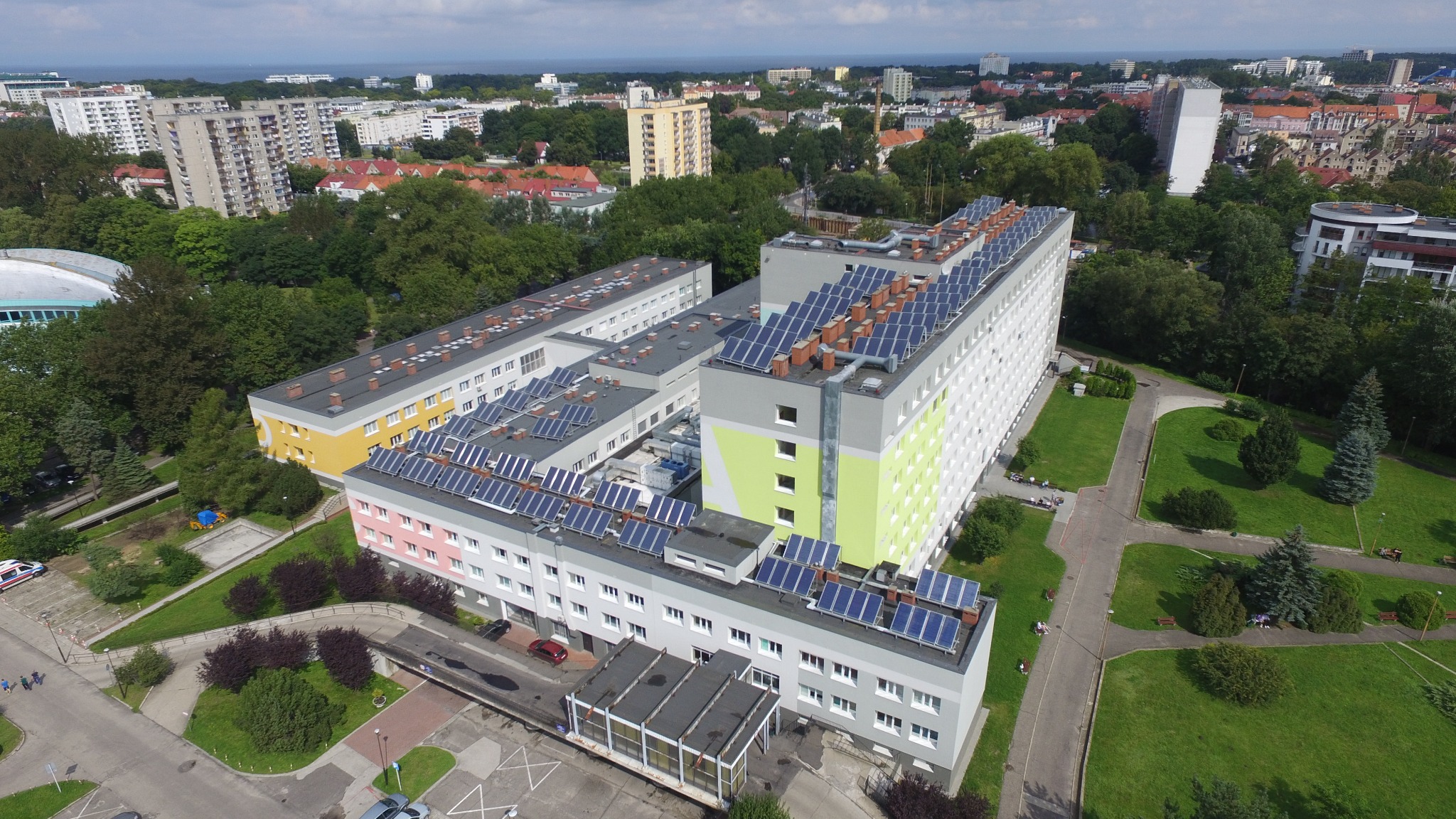 Widok z lotu ptaka na budynek z panelami słonecznymi na dachu, otoczony zielenią i zabudową miejską w tle.