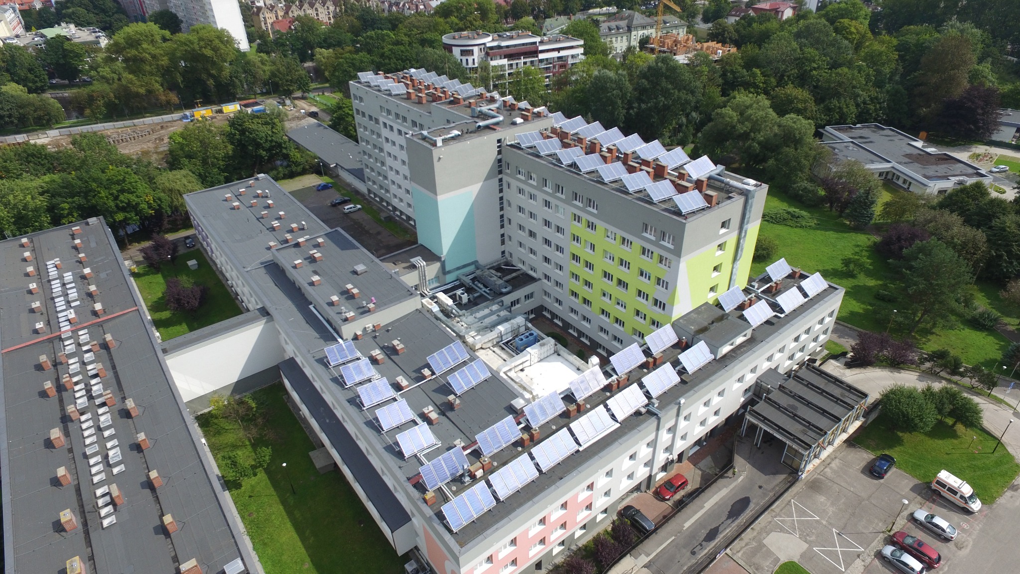 Widok z lotu ptaka na budynek komercyjny z panelami słonecznymi na dachu, otoczony zielenią i parkingiem z kilkoma samochodami.