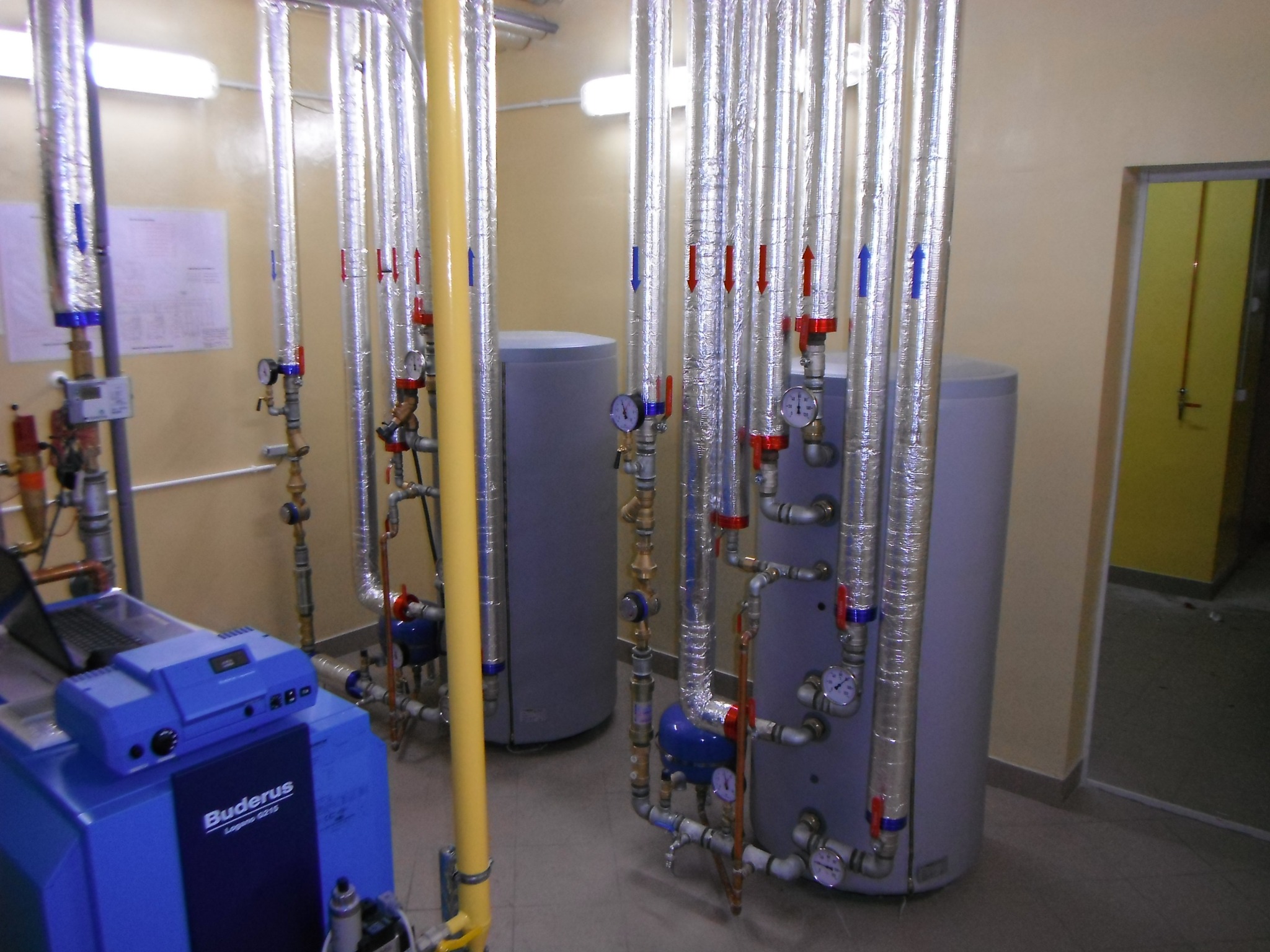 Złożona instalacja grzewcza z dwoma zbiornikami, licznymi rurami w srebrnej izolacji, manometrami i niebieskim kotłem Buderus Logano G215 z podłączonym laptopem, widoczna w pomieszczeniu z beżowymi...