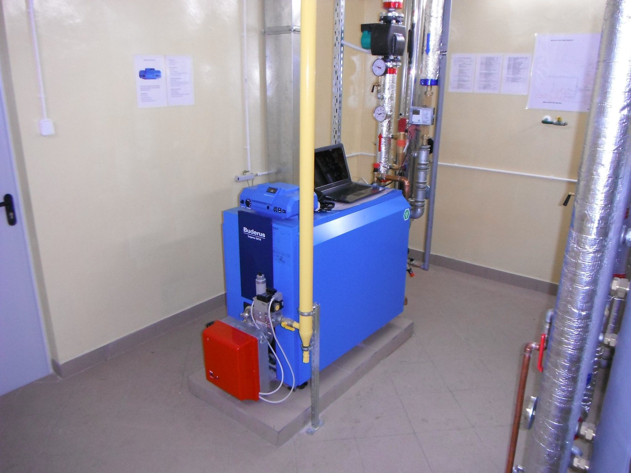 Niebieski piec Buderus z podłączonym laptopem, widoczne rury instalacji grzewczej w izolacji, czerwona obudowa palnika, żółta rura doprowadzająca gaz, dokumentacja techniczna na ścianie.