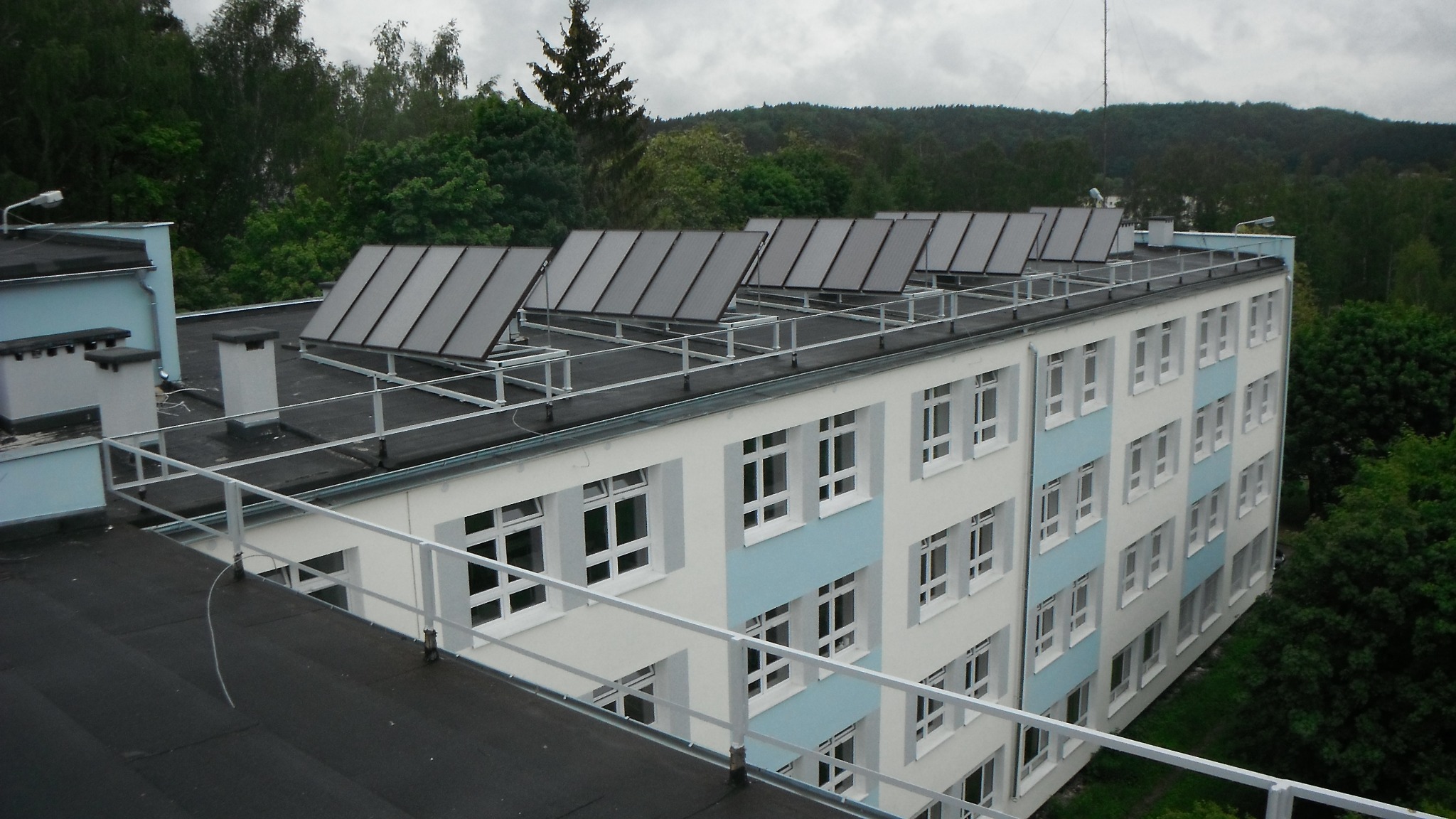 Rząd paneli słonecznych na dachu budynku z białymi i błękitnymi akcentami, widok z góry na tle zieleni.