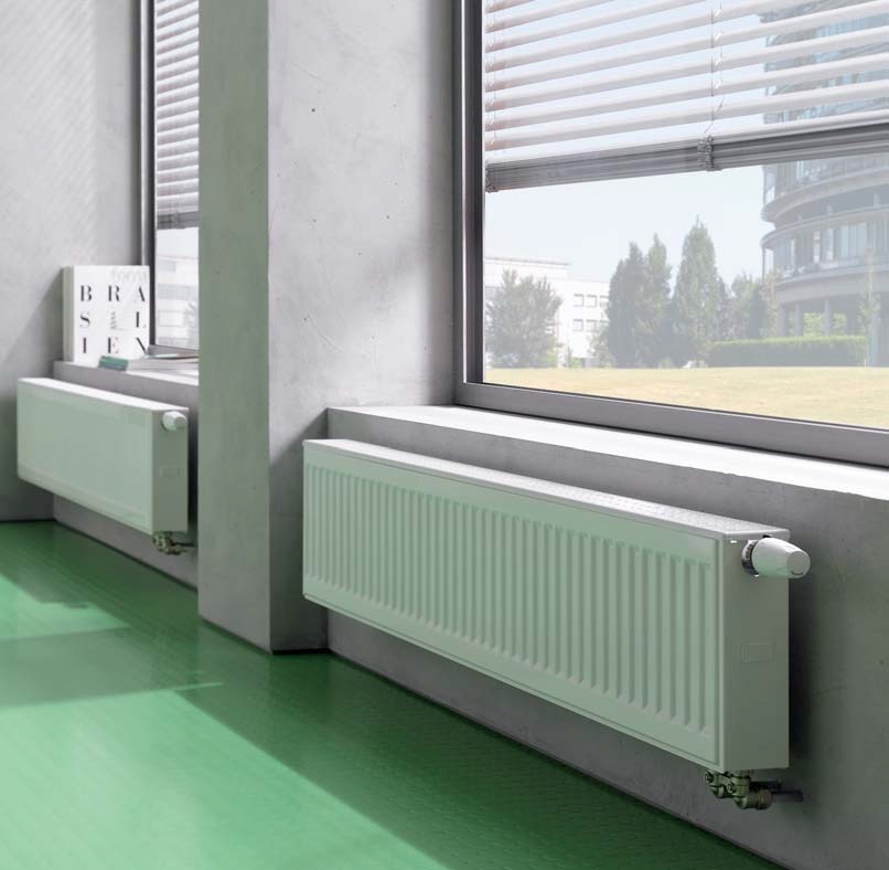 Dwa białe grzejniki panelowe zamontowane pod oknami na tle zielonej podłogi i surowych, betonowych ścian.