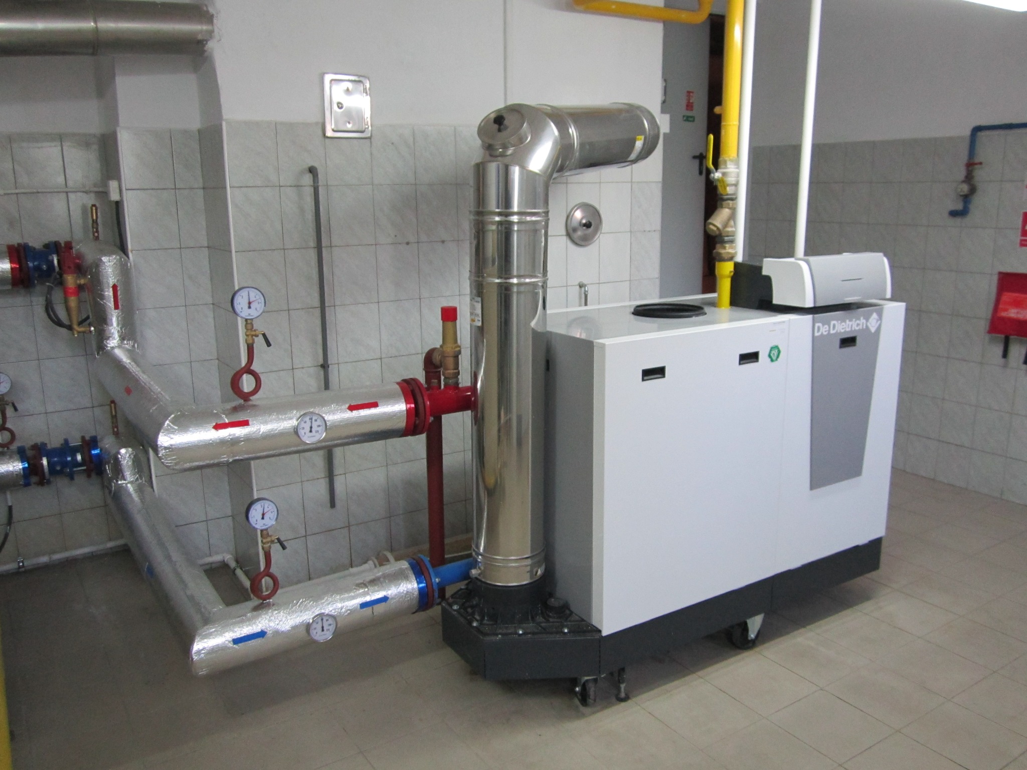 Nowoczesny, biały piec grzewczy De Dietrich z chromowanym kominem i izolowanymi rurami z manometrami w kotłowni wyłożonej kafelkami.